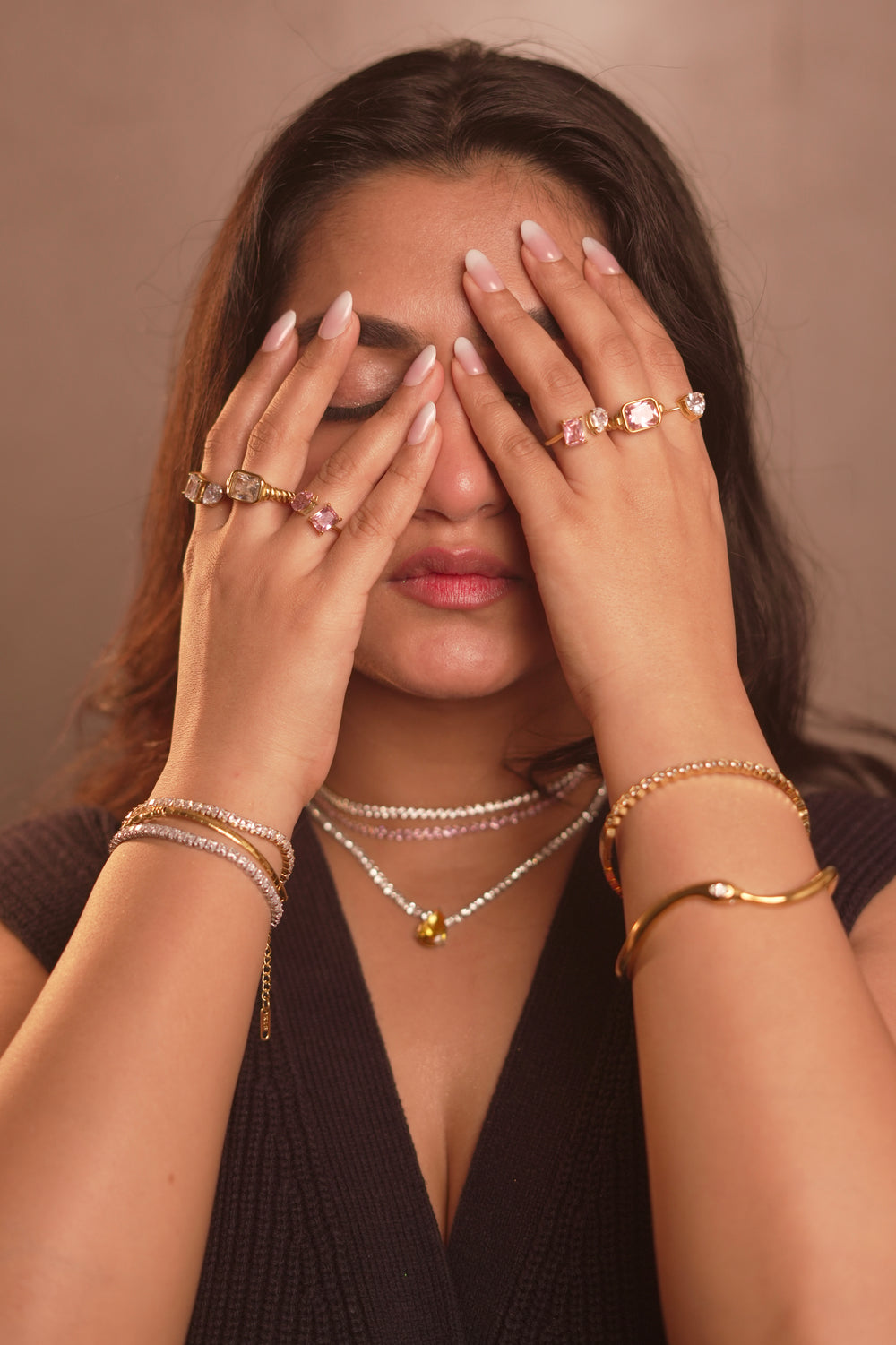 Woman covering her face with her hands, wearing multiple pieces of jewelry against a neutral background